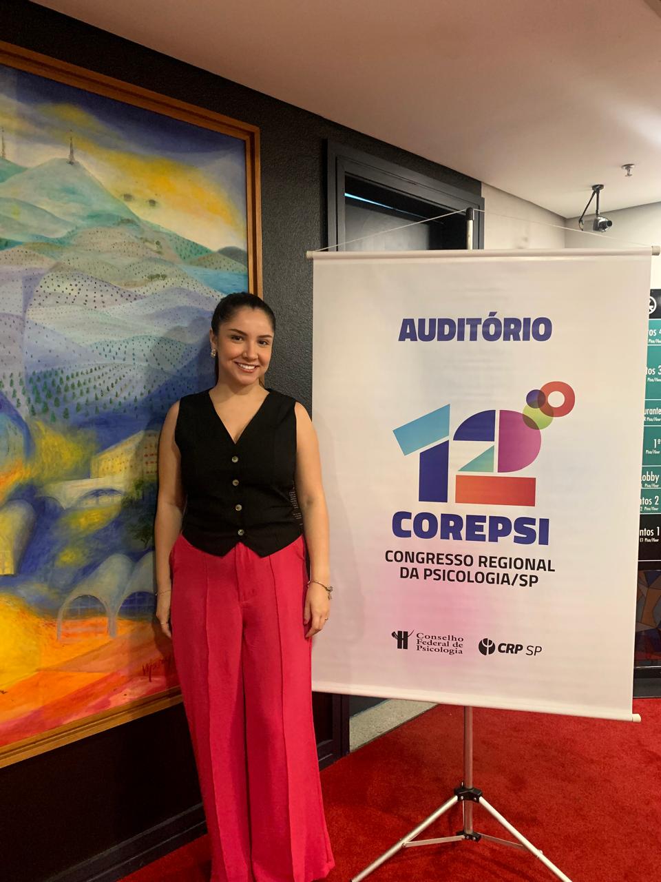 Ana Beatriz ao lado do banner do 12º COREPSI, Congresso Regional da Psicologia de São Paulo